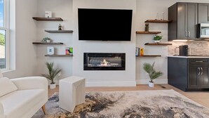 Uma sala de estar moderna com lareira, uma TV de tela plana na parede e uma cozinha com armários escuros e micro-ondas.