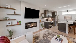 Uma sala de estar moderna com lareira, televisão de tela plana e uma cozinha com armários escuros e eletrodomésticos de aço inoxidável.