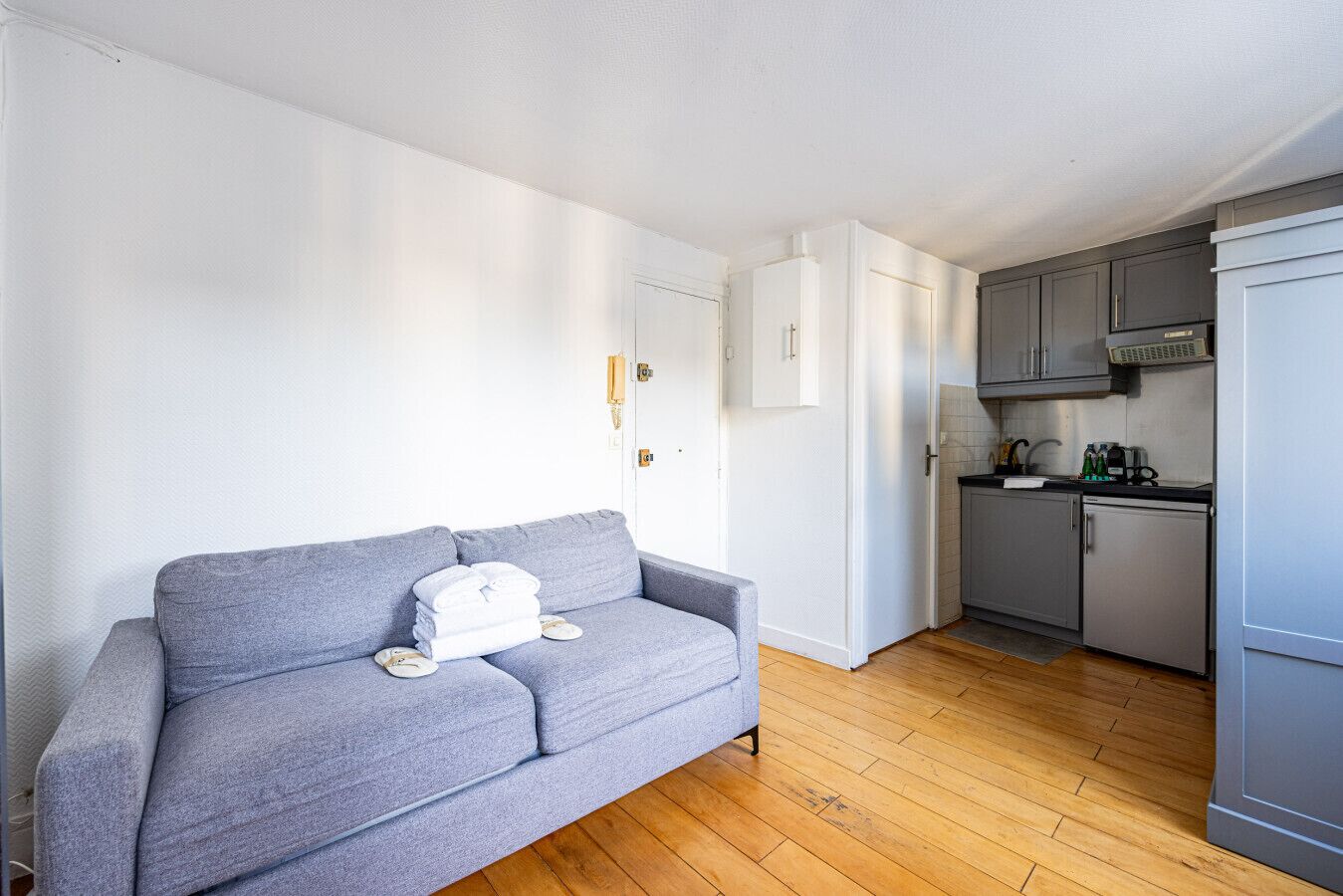 Un espacio habitable compacto con un sofá gris, una pequeña cocina y una puerta blanca.