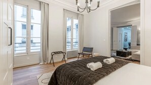 A modern hotel room with a large bed, two windows with curtains, a small seating area, and a chandelier. - 21.#Opera#Louvre#Paris02#2bedrooms#AC (Paris)