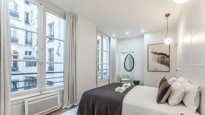 A hotel room with a bed, a chair, a round mirror, and a painting on the wall. - 21.#Opera#Louvre#Paris02#2bedrooms#AC (Paris)