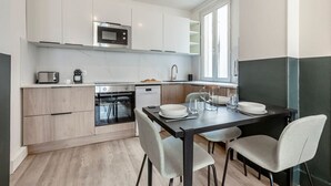 Une cuisine moderne avec des meubles blancs, un micro-ondes encastré, un four et un lave-vaisselle. On y trouve une table à manger avec des chaises et une fenêtre qui laisse entrer la lumière naturelle.