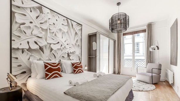 Une chambre moderne avec un grand lit, des panneaux décoratifs sur les murs, un luminaire suspendu et une vue sur la ville par la fenêtre.