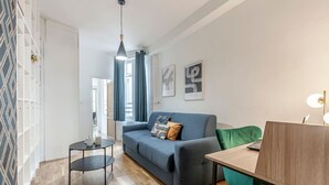 Un salon moderne avec un canapé, une petite table et un fauteuil. Il y a une fenêtre avec des rideaux, un luminaire suspendu et des œuvres d’art accrochées au mur.