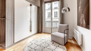 Une chambre moderne dotée d’une grande porte coulissante, d’un fauteuil gris, d’un tapis blanc et d’une fenêtre donnant sur des immeubles.