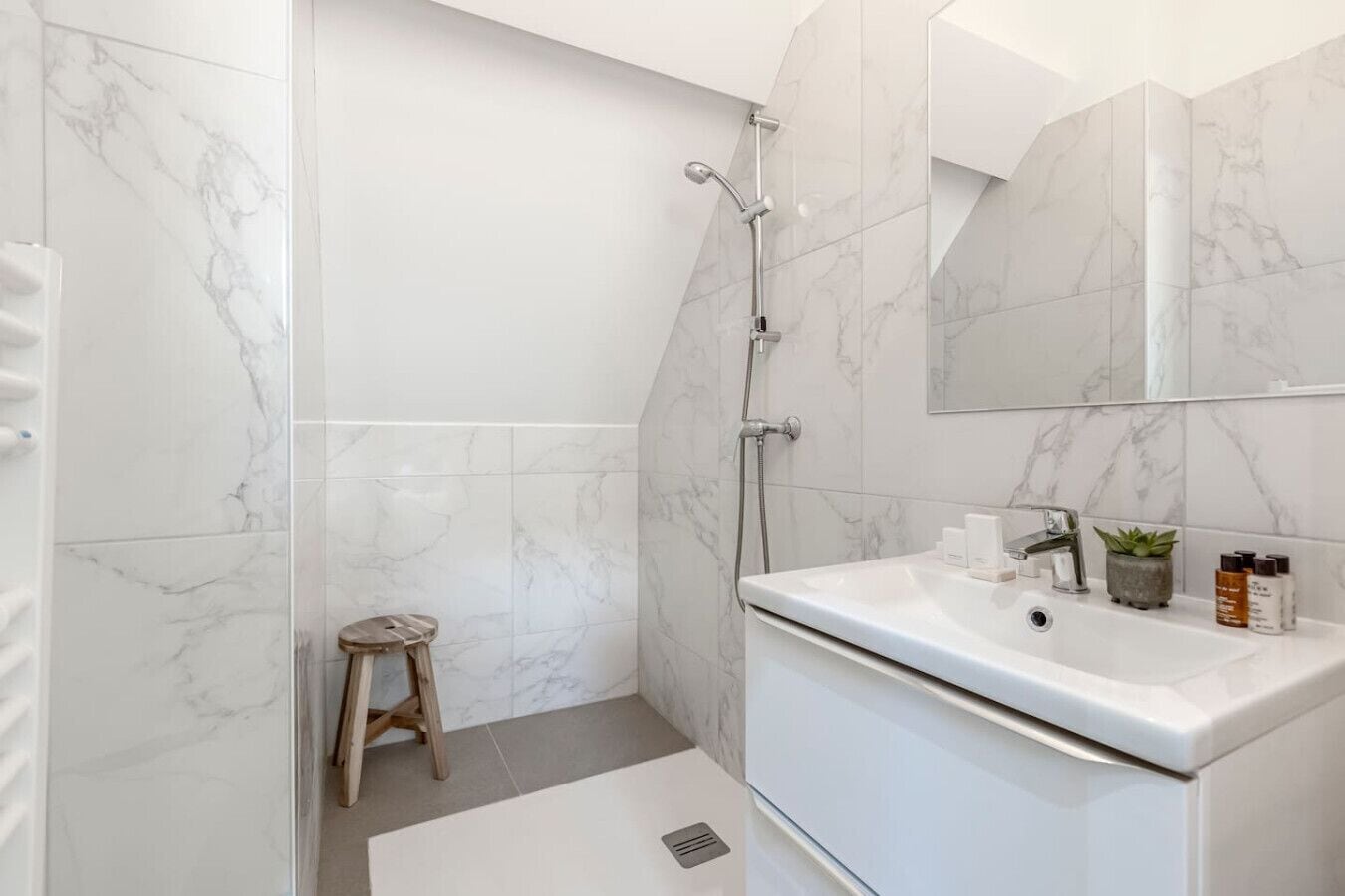 Een moderne badkamer met een witte marmeren douche, een houten krukje, een wit wastafelmeubel met toiletartikelen en een grote spiegel.