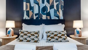A hotel room with a bed, two pillows with geometric patterns, two bedside tables with lamps and water bottles, and a large abstract artwork on the wall.