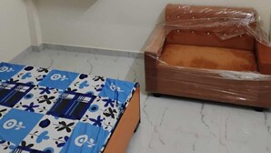 A room with a bed, a small sofa, a wall-mounted shelf, and a tiled floor. - Geeta Teerth Guest House (Faizabad)