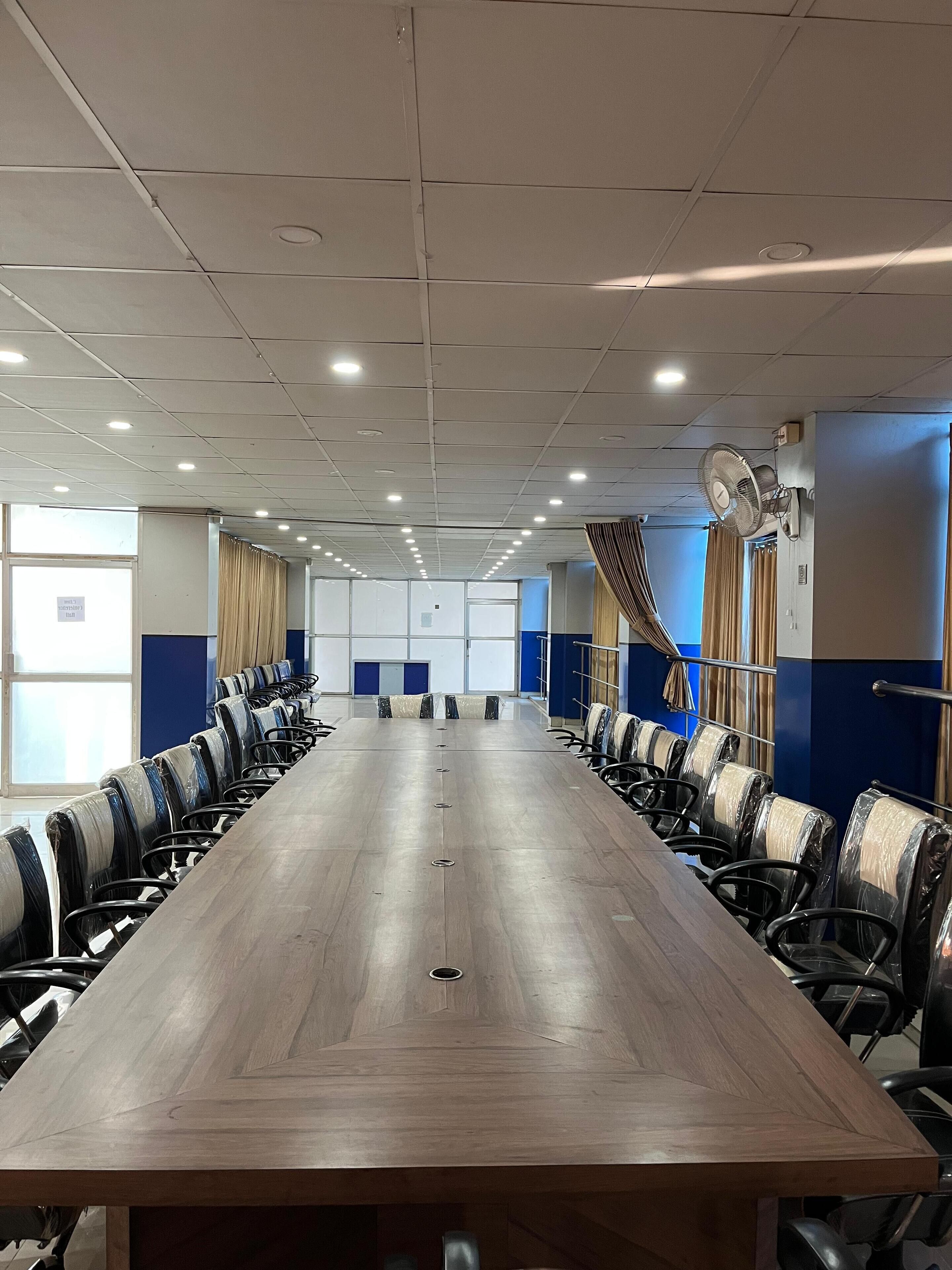 Un salón de conferencias con una larga mesa, sillas, luces de techo y un ventilador.
