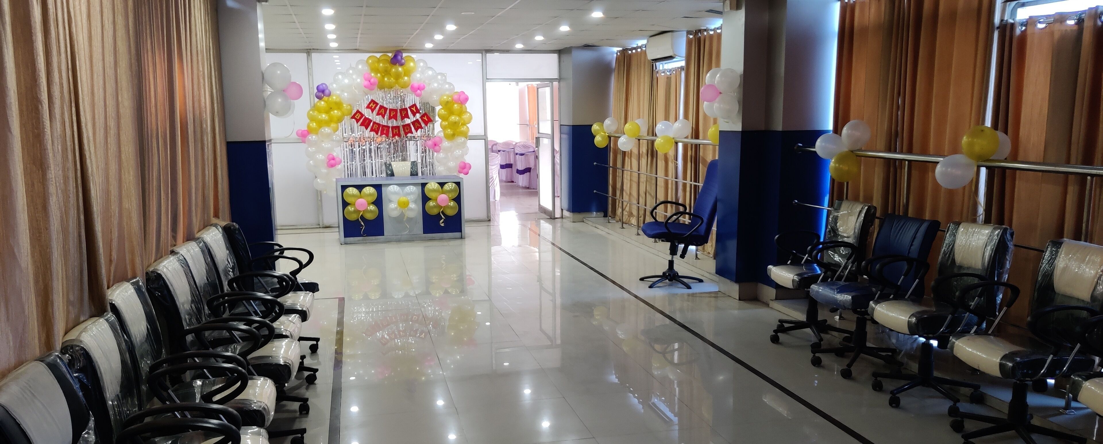 Un pasillo amplio con filas de sillas de oficina negras a ambos lados, que conduce a una zona de recepción decorada con globos y un cartel que dice 'Feliz Cumpleaños'.