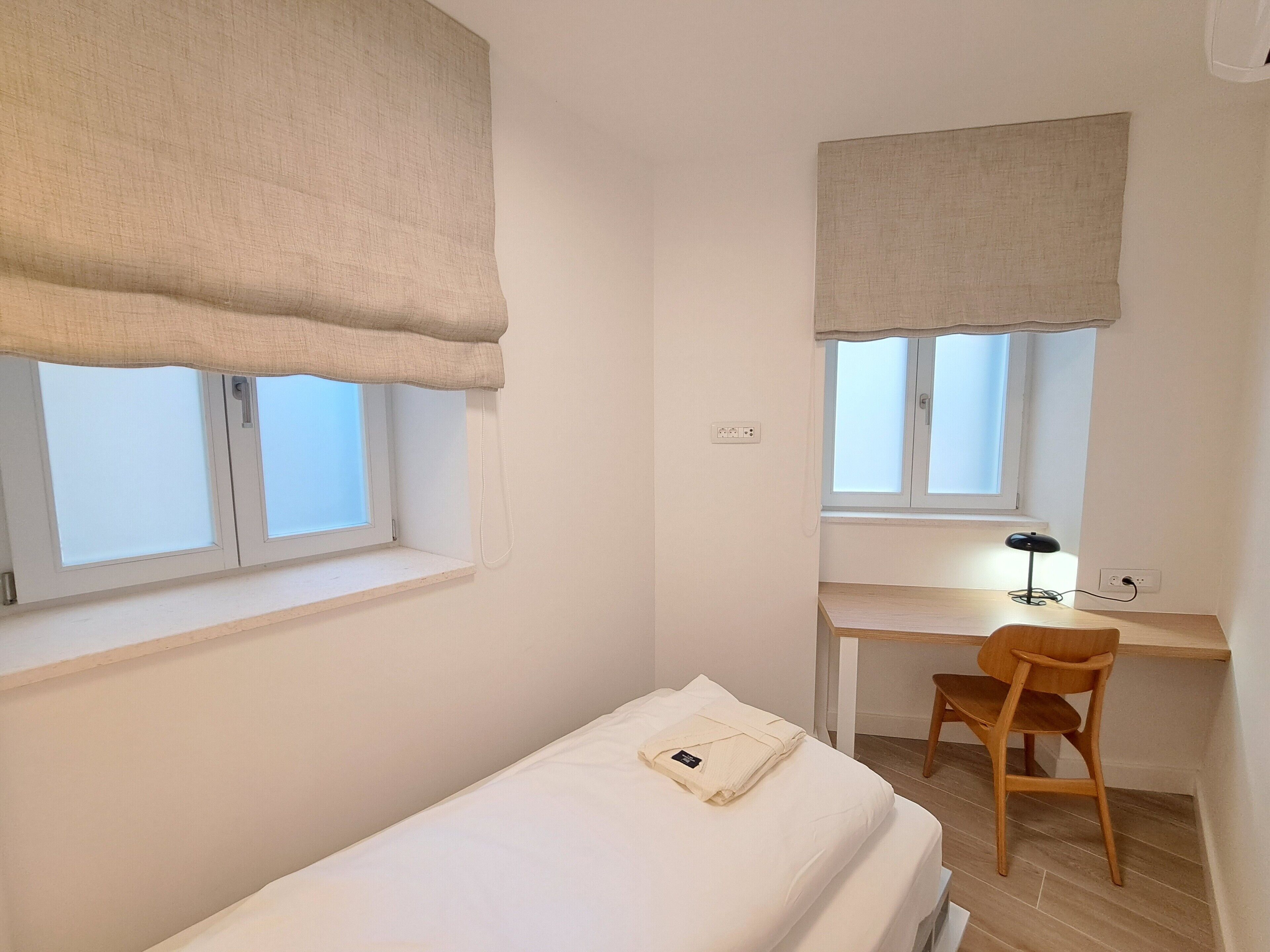 Habitació petita i senzilla amb un llit individual, un escriptori i una cadira. Hi ha dues finestres amb cortines, una a cada costat de la habitació.