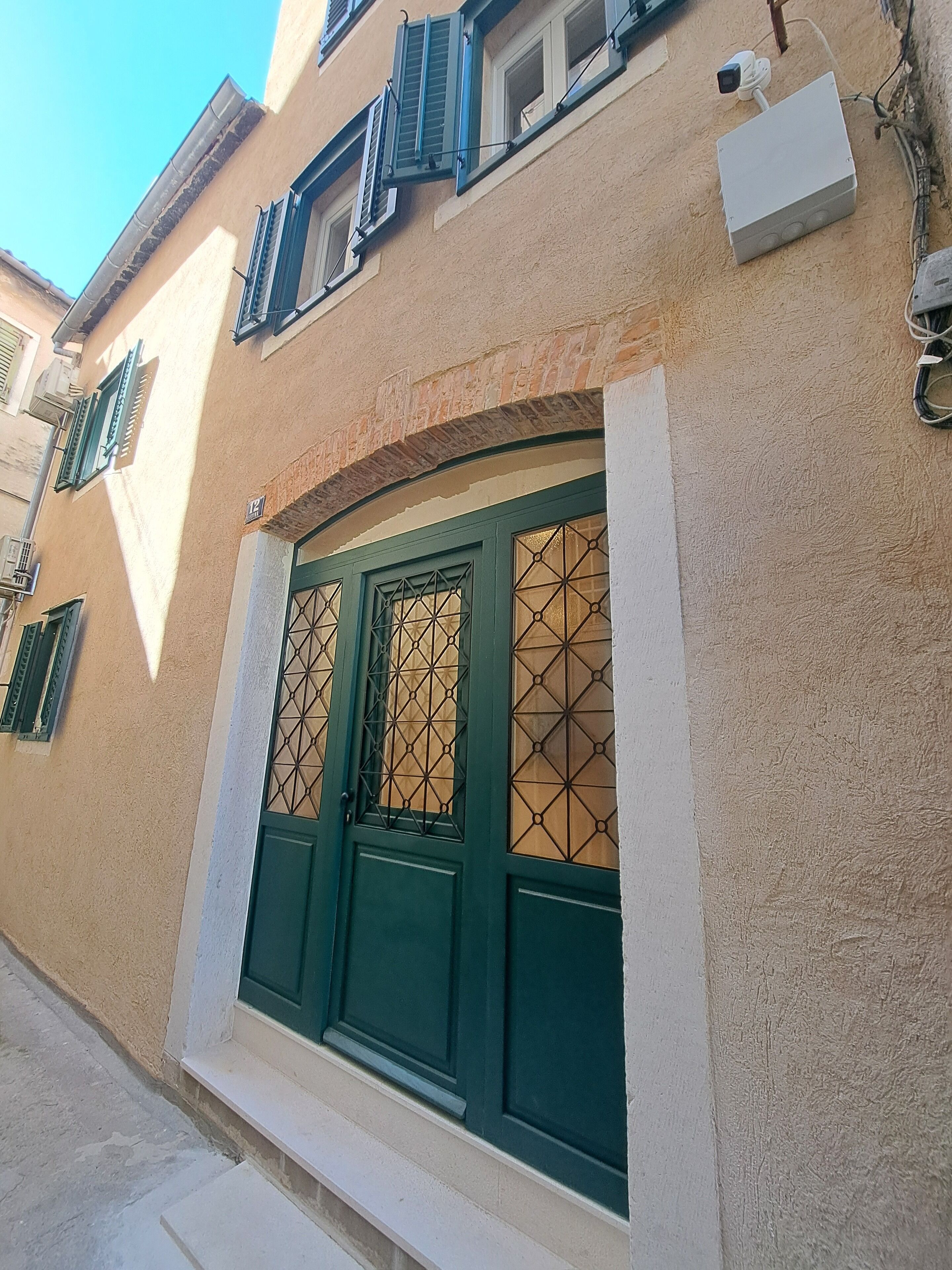 Un edificio con fachada color beige, contraventanas verdes y un amplio ventanal de arco con un diseño geométrico.