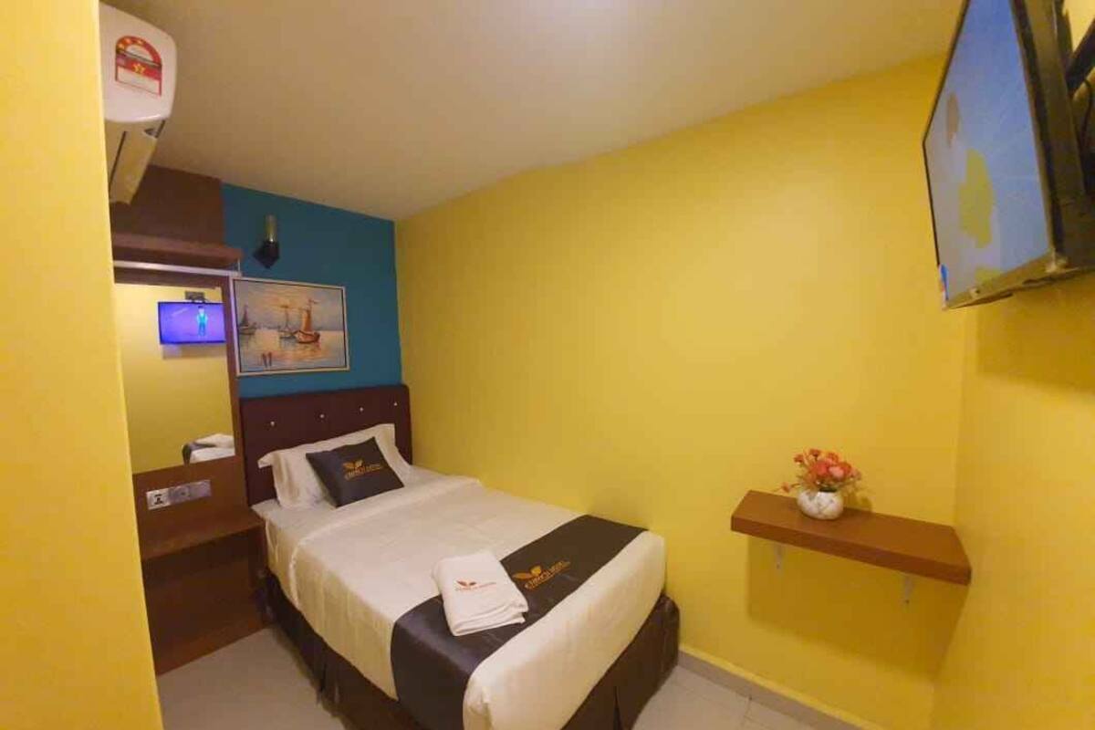 Une chambre d’hôtel avec un lit, un petit bureau, une télévision et une étagère fixée au mur, ornée de fleurs.