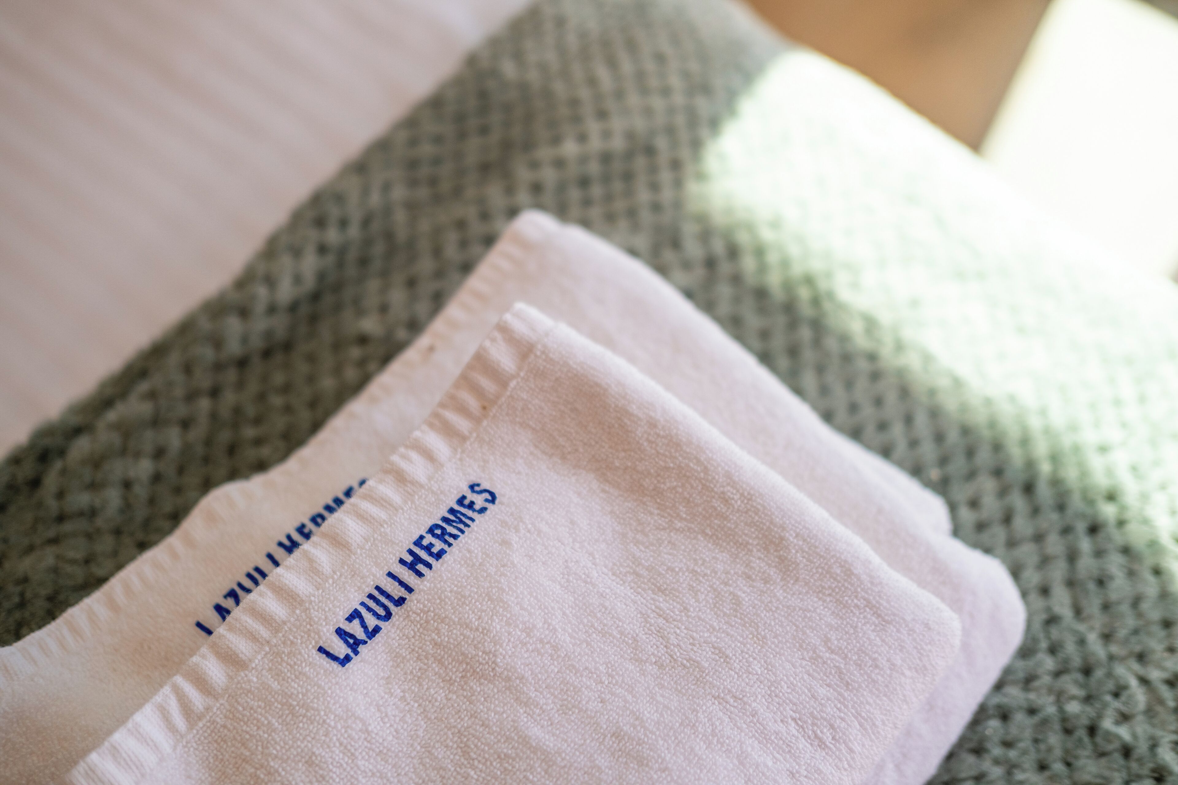 Une serviette blanche pliée, ornée de l’inscription « LAZULI HERMES », est posée sur une surface texturée.