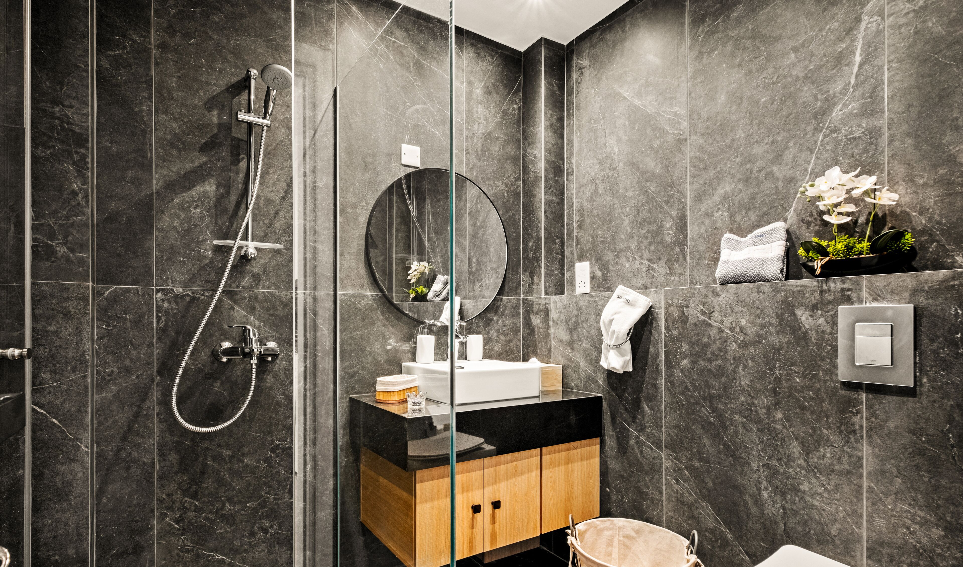 A modern bathroom with dark marble tiles, a glass-enclosed shower, a round mirror, a wooden vanity, and a small plant on the counter.