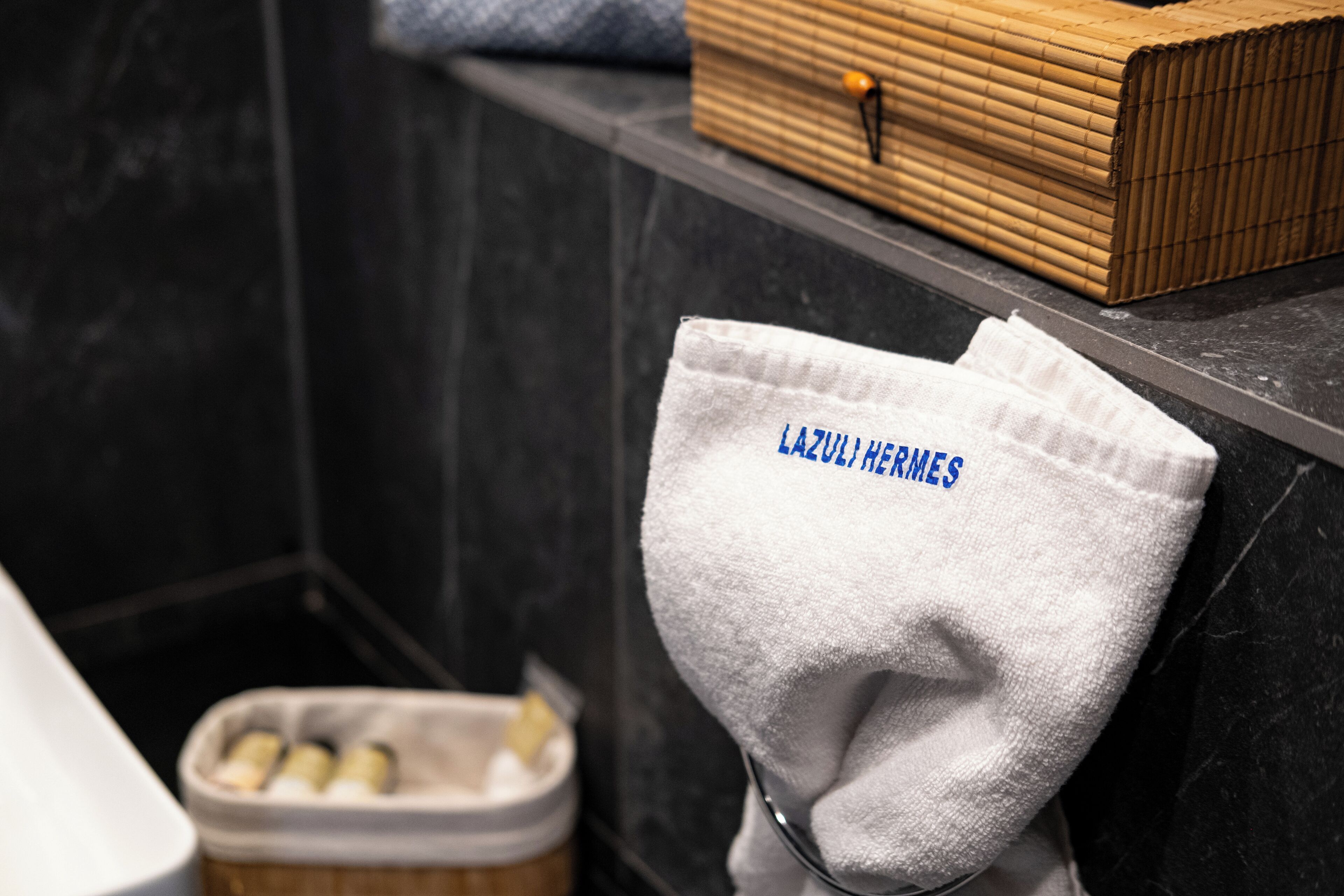 Un baño con un perchero que sostiene una toalla blanca con el bordado "LAZURIERI HERMES", una caja de madera en el estante y un recipiente con artículos de aseo.