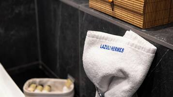 Un baño con un perchero que sostiene una toalla blanca con el bordado "LAZURIERI HERMES", una caja de madera en el estante y un recipiente con artículos de aseo.