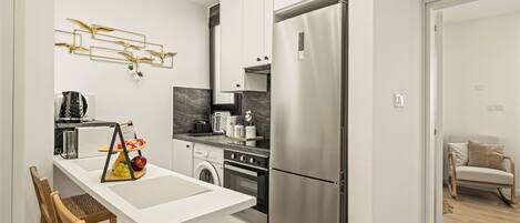 A modern kitchen with a stainless steel refrigerator, a built-in oven, and a washing machine. There is a dining table with chairs and a decorative wall piece above it.