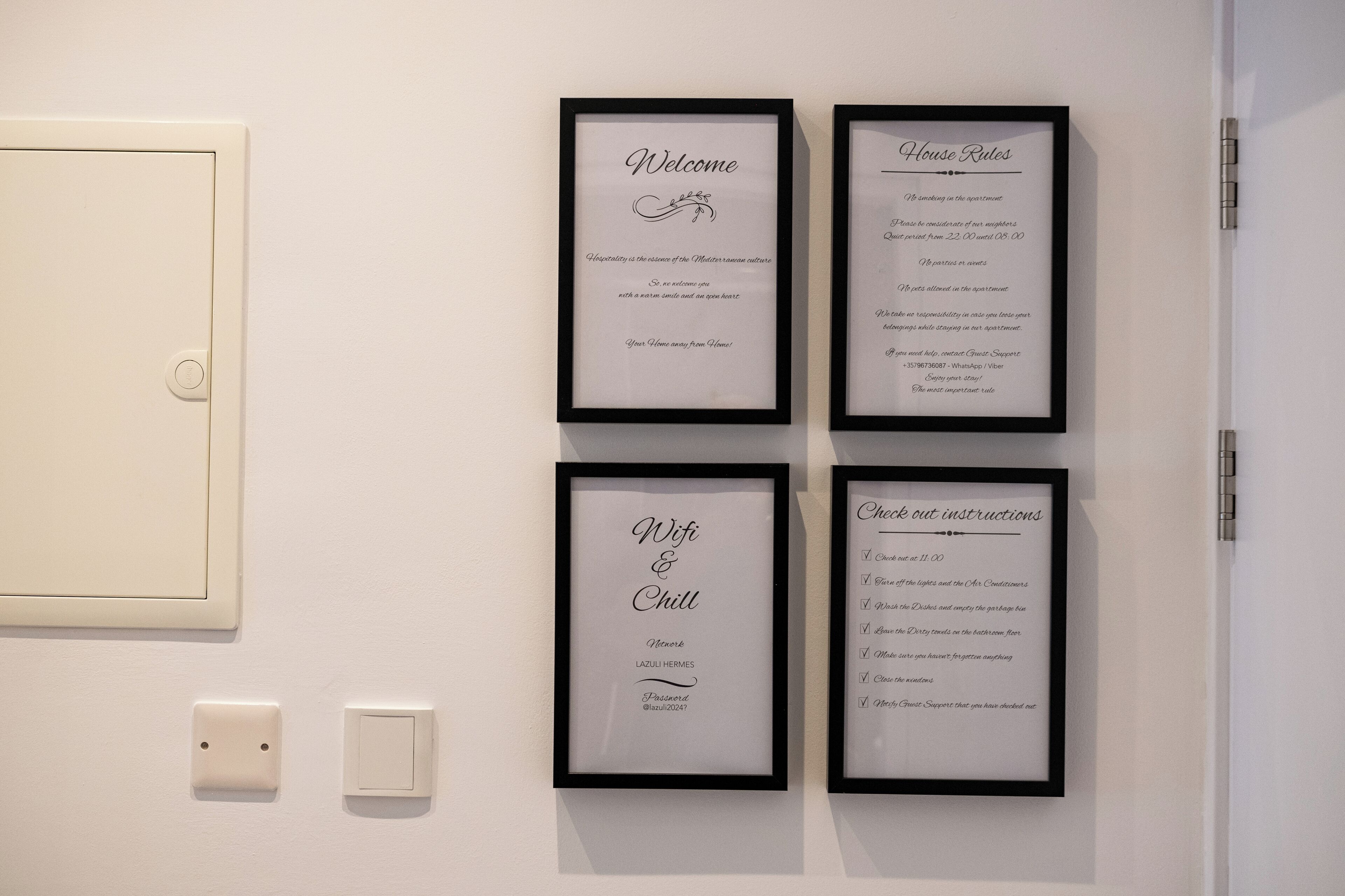 Four framed welcome and house rules signs on a wall, two light switches below them, and a door to the left.