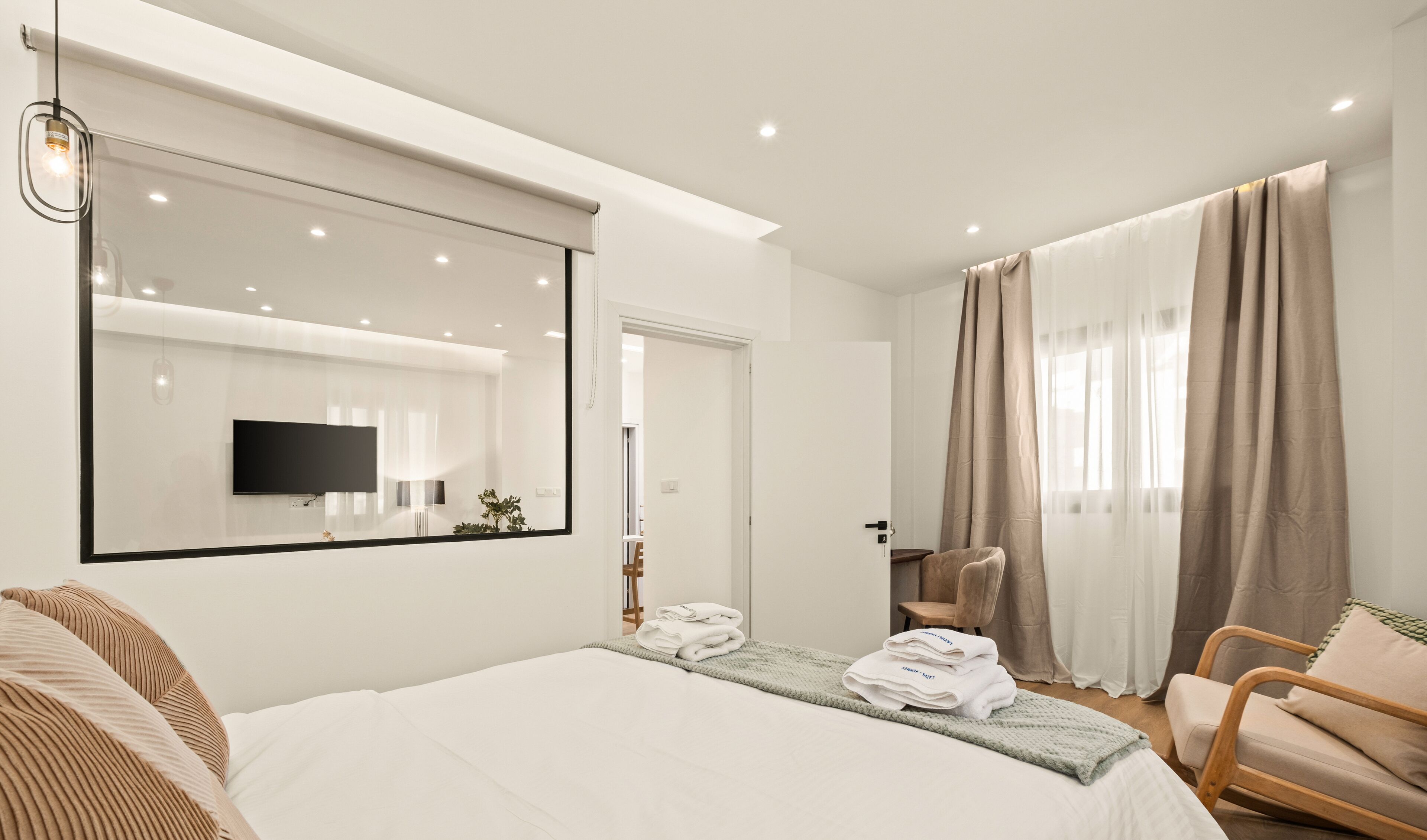 Une chambre d’hôtel moderne avec un grand lit, un téléviseur fixé au mur, une chaise en bois et une grande fenêtre avec des rideaux.