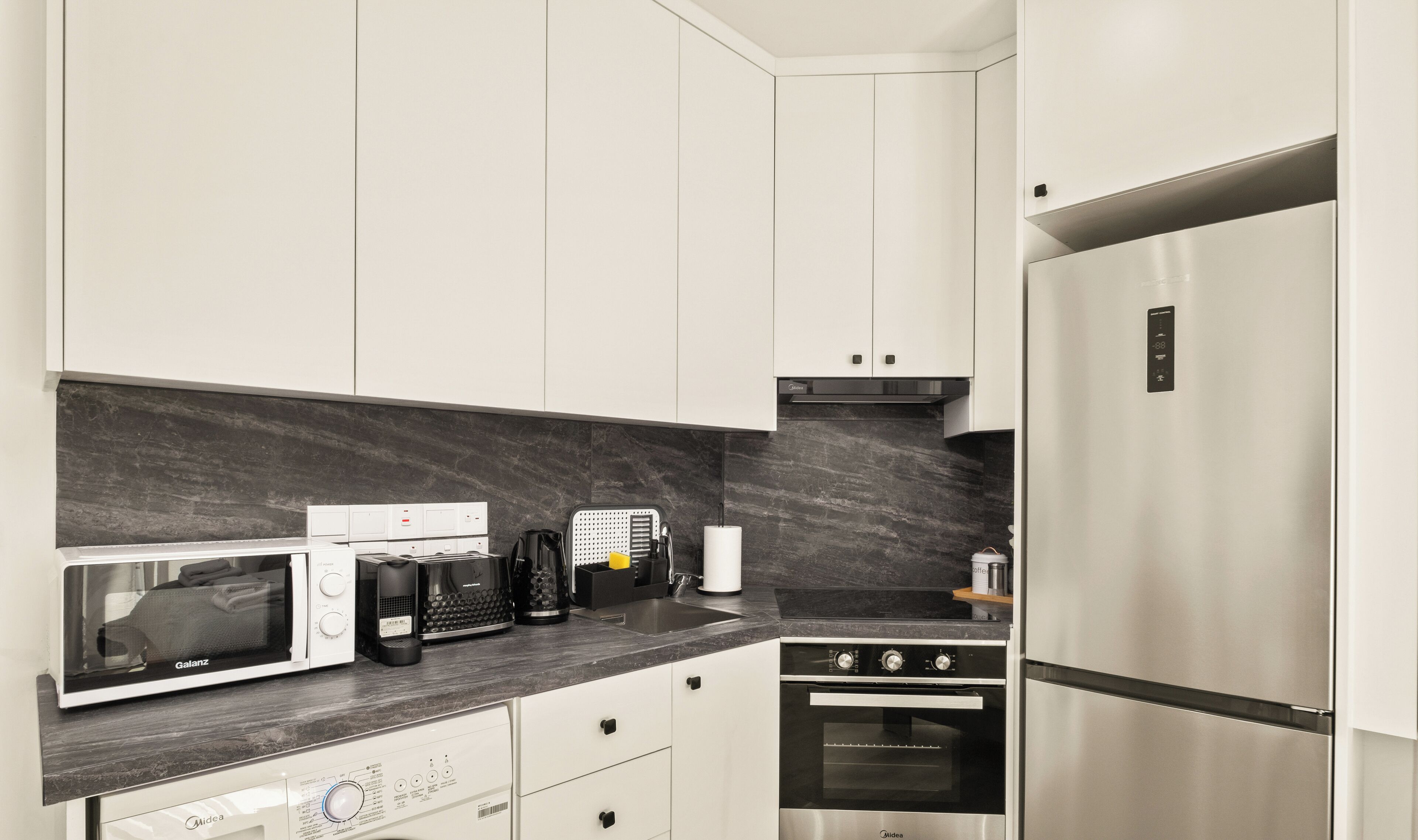 Una cocina moderna con armarios blancos, encimera negra, horno integrado, refrigerador de acero inoxidable y microondas.