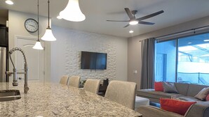 Eine moderne Küche mit Granitarbeitsplatten, Pendelleuchten, einem eingebauten Ofen und einem Essbereich mit grauem Sofa und roten Kissen.