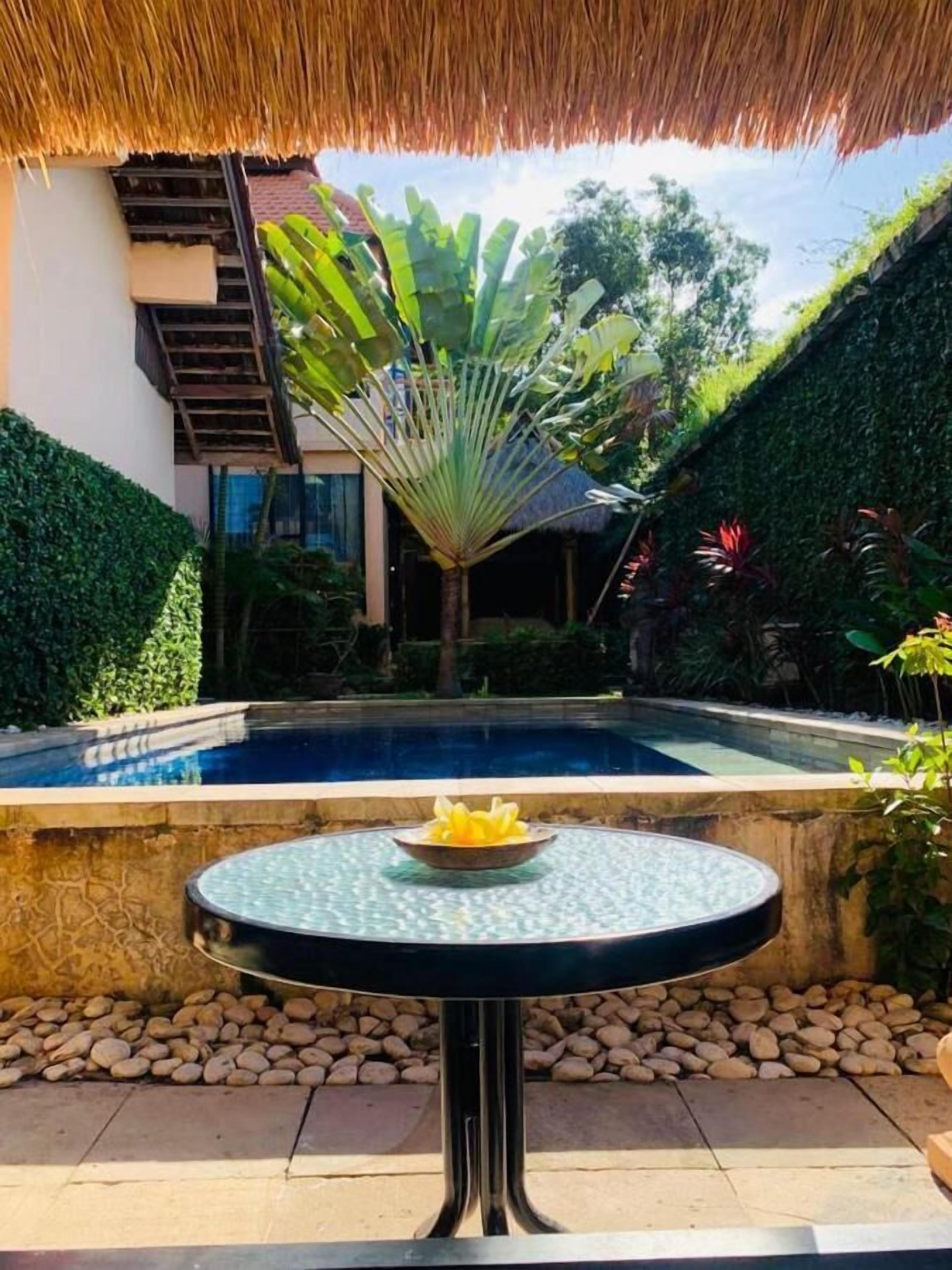 A poolside area with a round table, a thatched roof, and lush greenery.