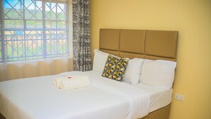 Un lit bien fait, avec des draps blancs et une serviette enroulée, un coussin à motifs et une tête de lit adossée à un mur jaune.
