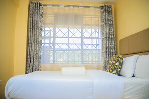 A bed with white bedding, a patterned curtain, and a window with a grid design. - Lux Suites J'Melots Apartments Kisumu (Kisumu)