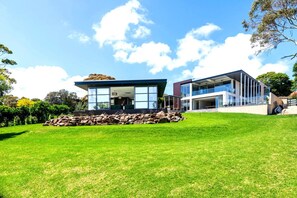 A modern house with a large glass facade, a stone wall in front, and a well-maintained lawn. - Panoramic Bay Views Mt Martha (Mount Martha)