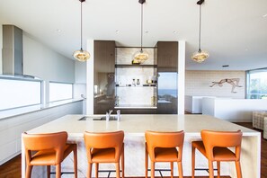 A modern kitchen with a central island, three bar stools, pendant lighting, and a view of the outdoors. - Panoramic Bay Views Mt Martha (Mount Martha)