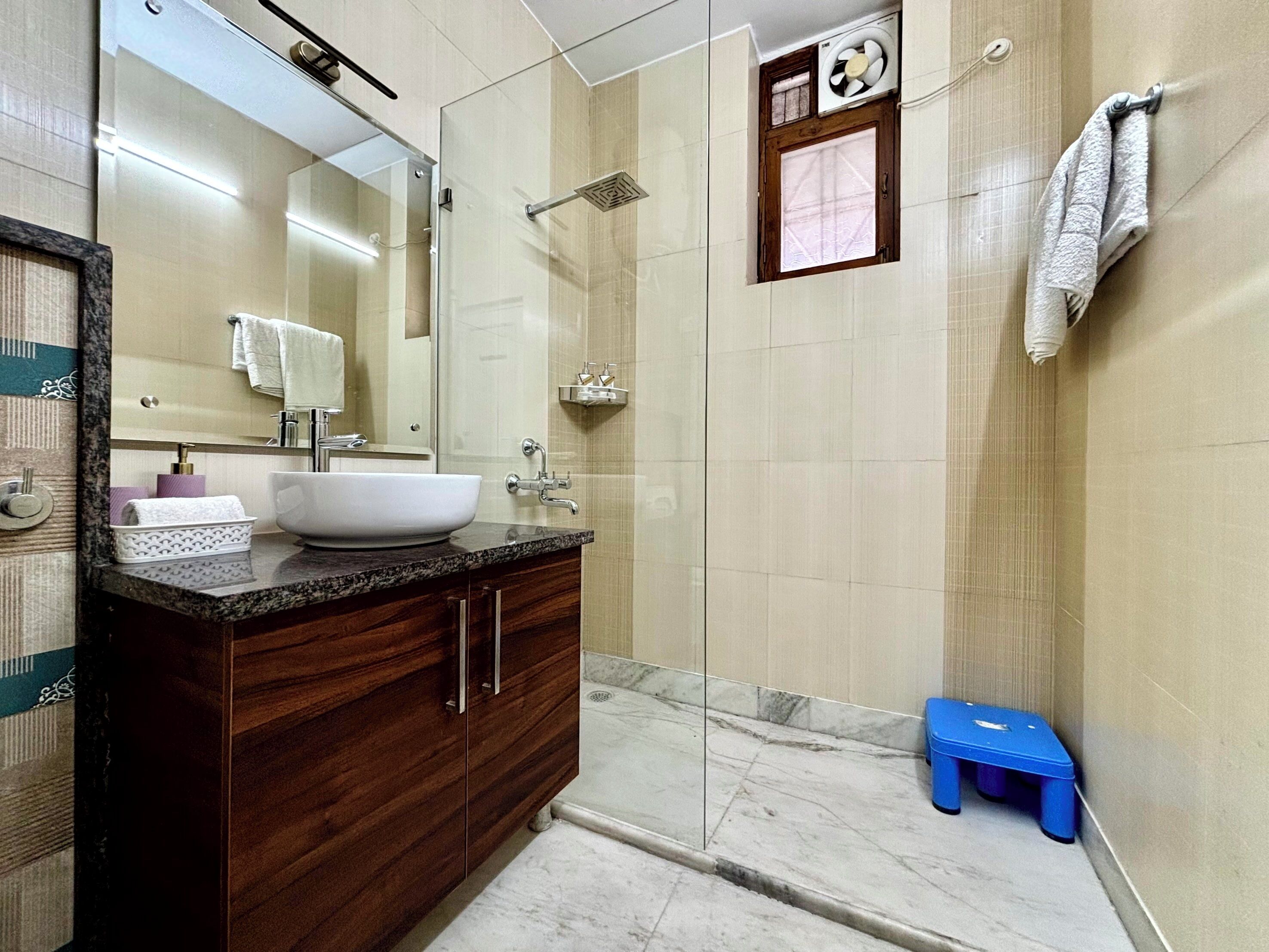 Un baño moderno con un espejo grande, un lavamanos blanco, una ducha y un taburete azul.