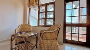 Ein Zimmer mit zwei Korbstühlen, einem kleinen Tisch und einem Fenster mit Blick auf Bäume und Gebäude.