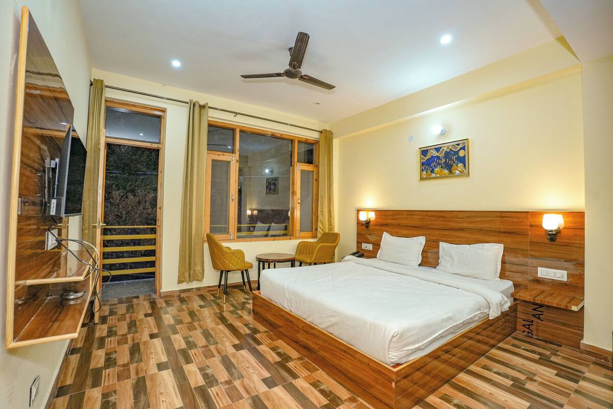 A hotel room with a bed, two chairs, a small table, a ceiling fan, and a painting on the wall.