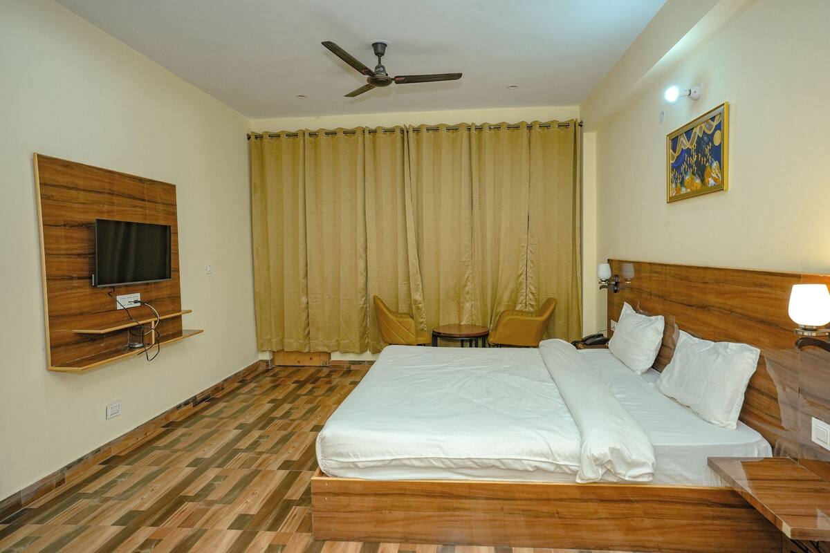 A hotel room with a wooden floor, a bed with white linens, a ceiling fan, a TV mounted on the wall, a table with chairs, and a painting on the wall.