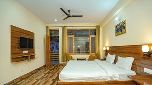 Habitación de hotel con una cama grande, un televisor montado en la pared, un ventilador de techo y una ventana con cortinas.