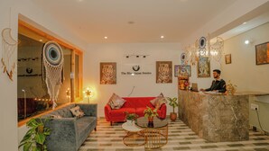 Un vestíbulo de hotel con una mesa de recepción, un sofá rojo, un sofá gris, una mesita de centro y plantas decorativas.