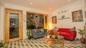 Una sala de estar con un sofá rojo, un sofá azul y una mesita de centro. Hay plantas, un atrapasueños y un cuadro de un tigre en la pared.