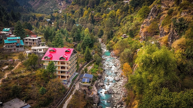 Un hotel de varios pisos con techo rojo, ubicado junto a un río de lecho rocoso, rodeado de un bosque denso y terreno montañoso.