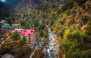 A multi-story hotel with a red roof, situated beside a river with a rocky bed, surrounded by dense forest and mountainous terrain. - The Himalayan Stories  (Manali)