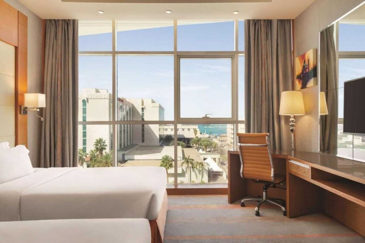 A hotel room with two beds, a desk, a chair, a TV, and large windows offering a city view.