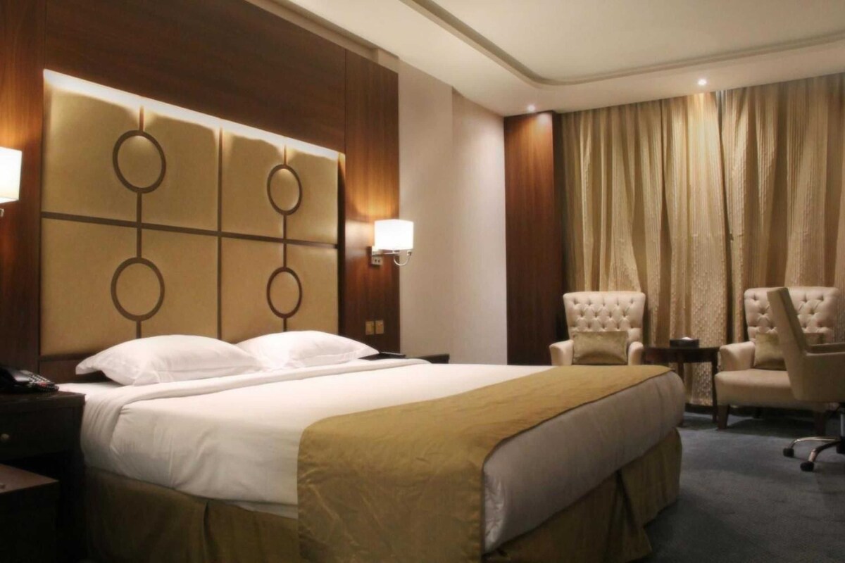A hotel room with a large bed, two bedside tables, a chair, a desk, and a curtain.