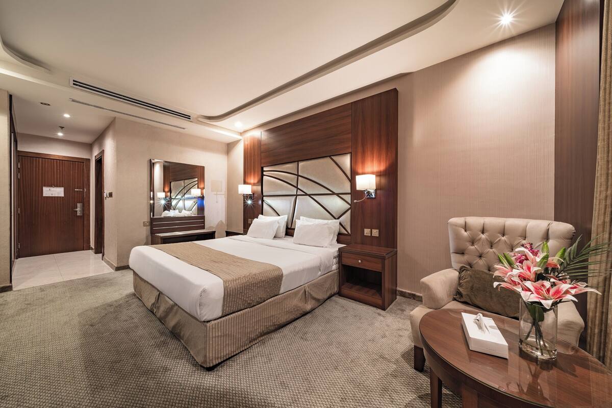 A hotel room with a large bed, a sitting area with a chair, a nightstand, and a vase of flowers.