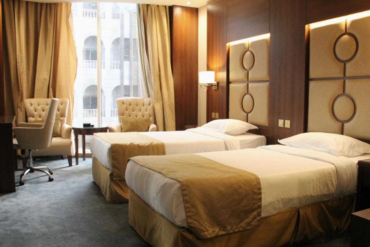 A hotel room with two beds, a desk, a chair, and a view of a building through large windows.