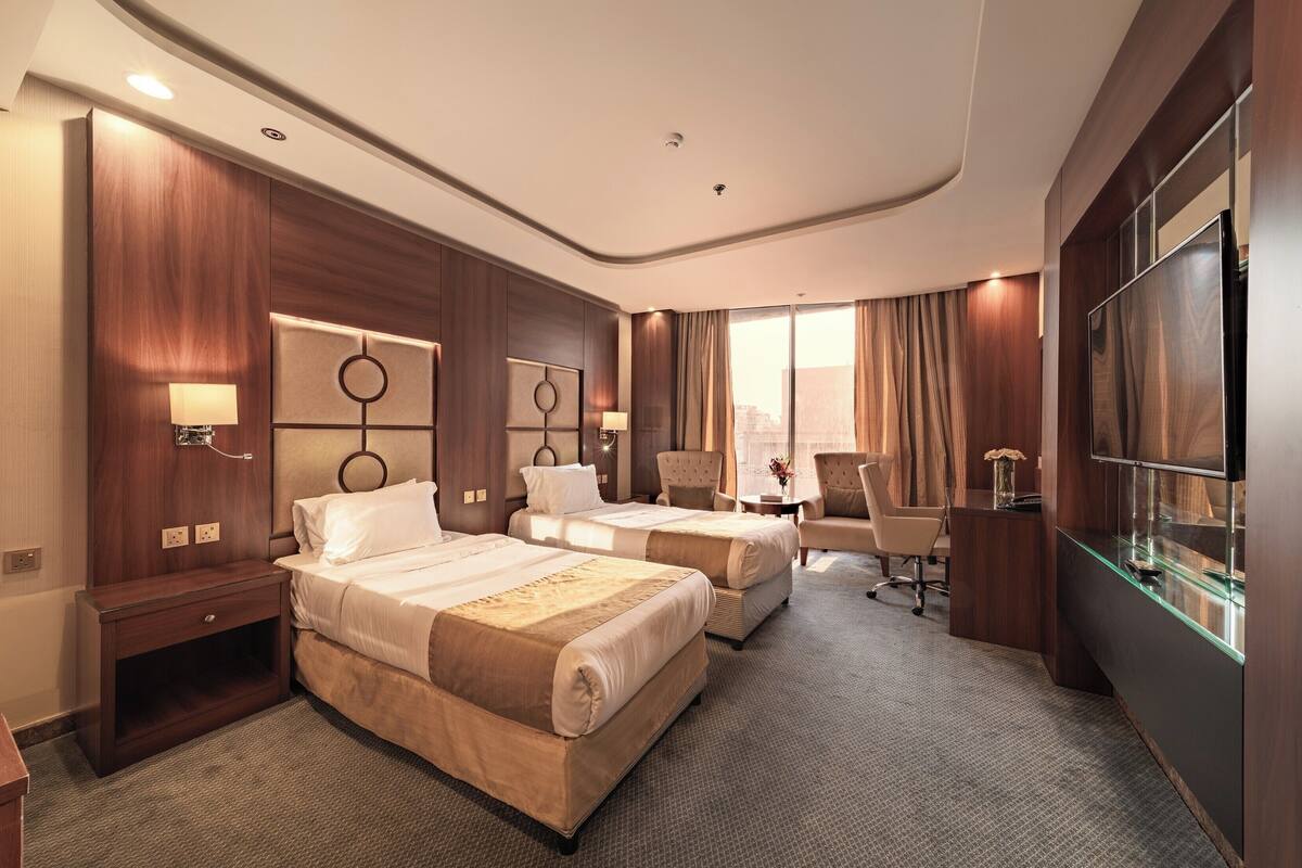 A hotel room with two beds, a desk, a chair, a TV, and a large window.