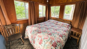 Une chambre située dans une cabane en bois, avec un grand lit, deux fenêtres, une chaise et une petite table.