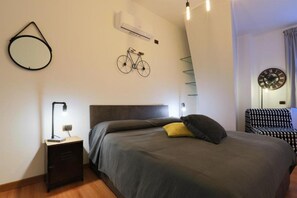 Un dormitorio con una cama, una mesita de noche, una obra de arte con motivos de bicicleta, un reloj y una silla estampada.