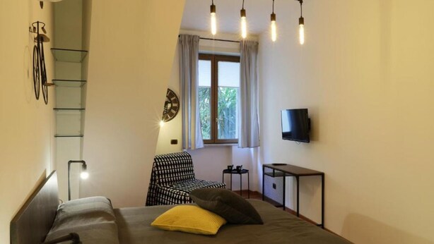 Un dormitorio con cama, una silla, televisor, ventana con cortinas y un soporte para bicicletas.