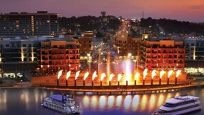 A waterfront hotel with a central fountain, boats docked, and surrounding buildings.