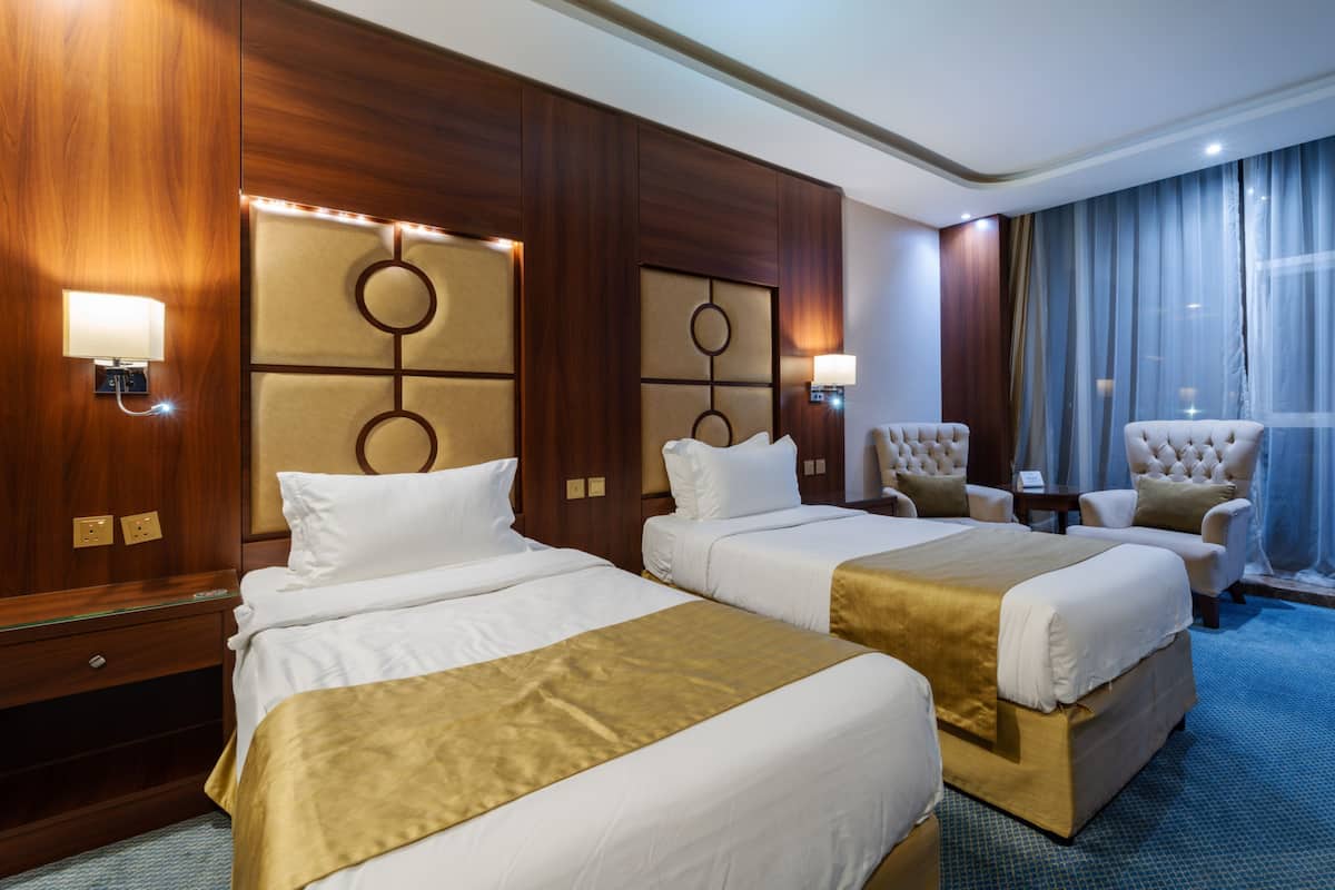 Ein Hotelzimmer mit zwei Betten, einem großen Fenster mit Vorhängen, einer Sitzecke mit einem Sessel und einer Wandlampe.